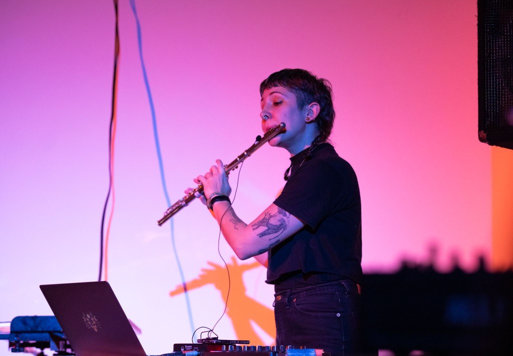 A non-binary person with light tan skin and dark hair in two braids, a short fringe, and an undercut to their temples, playing flute with a microphone attached and a cord running to the laptop in front of them. They are wearing a black t-shirt and black jeans and there is a wash of purple-pink-peach light washing over them and the white wall behind.